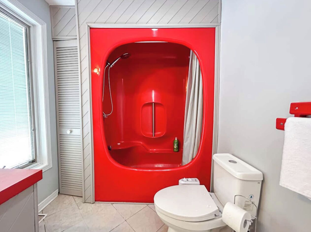A bathroom with a red bathtub, a white toilet, and a window with blinds.