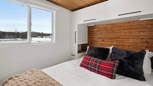 A bedroom with a large window, wooden ceiling, white walls, and a bed with multiple pillows.