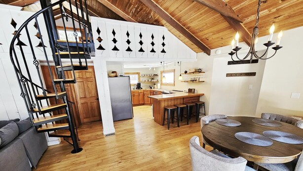 Amplia sala de estar con escalera de madera, una mesa de comedor con sillas y una cocina al fondo.