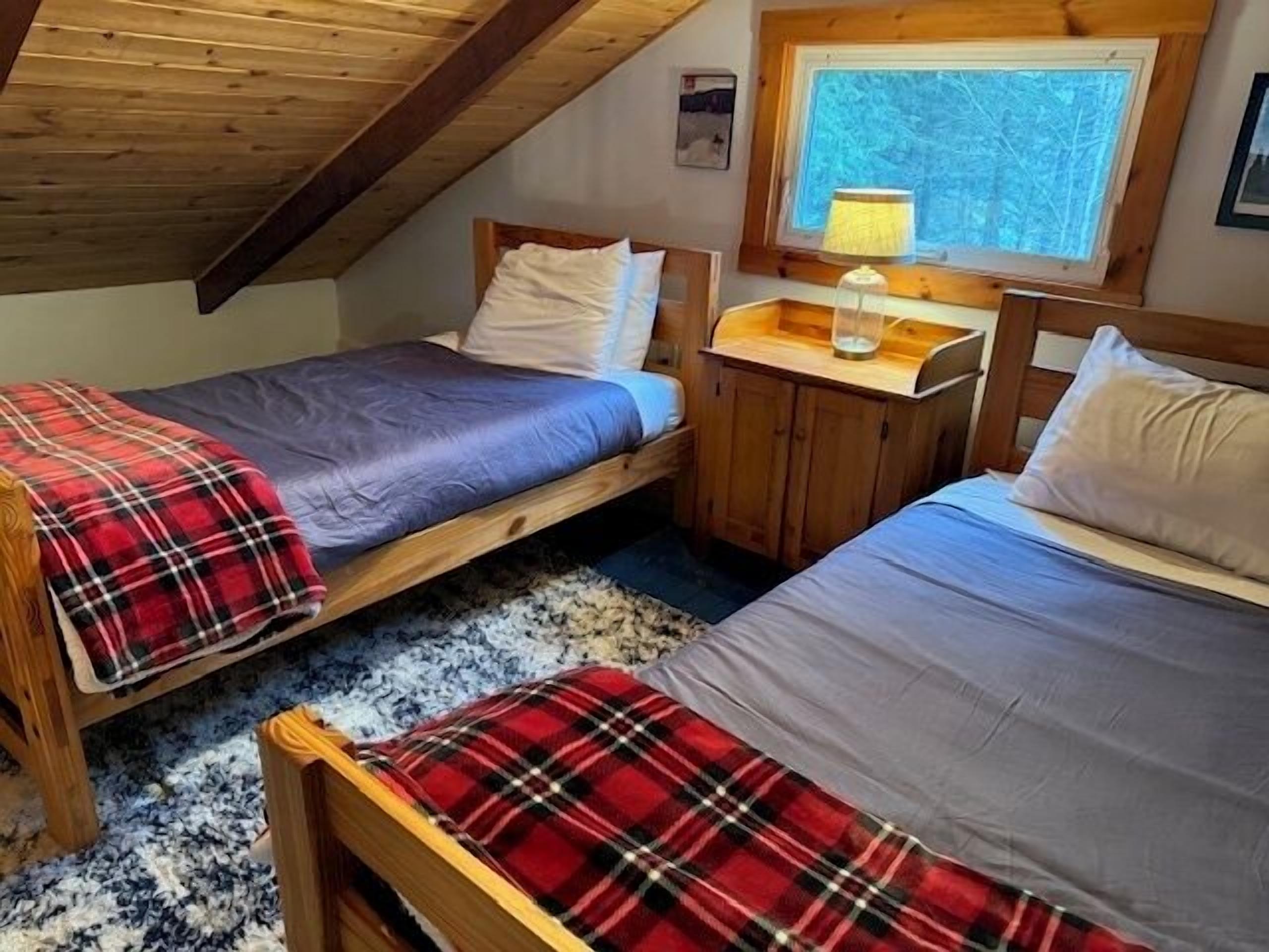 Une pièce chaleureuse avec deux lits en bois, une petite table en bois avec une lampe et une fenêtre donnant sur l’extérieur.