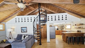 Un loft espacioso con techo alto, vigas de madera a la vista, una escalera espiral central, un sofá gris acogedor, una zona de cocina y un comedor con taburetes.