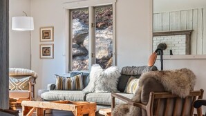 Ein gemütliches Wohnzimmer mit einer Couch, einem Holztisch und einem Sessel. An den Wänden hängen Bilderrahmen und durch das Fenster ist ein Blick auf Felsen möglich.