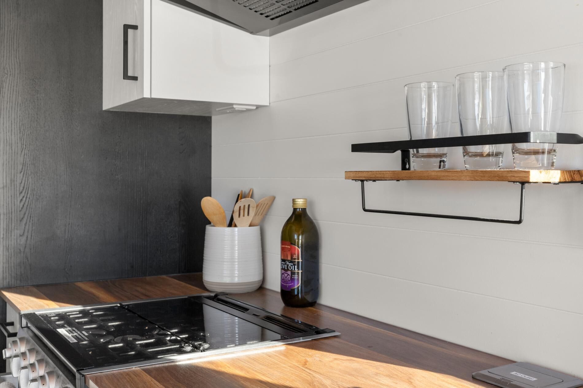 Una cocina moderna con una placa de inducción negra, un armario blanco y un estante de madera con copas y una botella de aceite de oliva.
