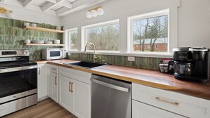 Une cuisine moderne avec des armoires blanches, une cuisinière en acier inoxydable, un four à micro-ondes, un évier et une machine à café Keurig. Trois fenêtres laissent entrer la lumière naturelle et offrent une vue sur les arbres et un bâtiment à l’extérieur.
