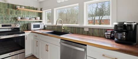 A modern kitchen with white cabinets, a stainless steel stove, a microwave, a sink, and a Keurig coffee maker. There are three windows providing natural light and a view of trees and a building outside.