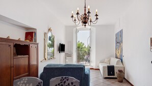 Une pièce comprenant un meuble en bois, un banc recouvert d’un tissu bleu, un canapé blanc, un lustre et une grande fenêtre donnant sur un paysage verdoyant.