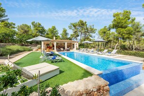 A swimming pool with lounge chairs and umbrellas, surrounded by greenery and a building with outdoor seating.