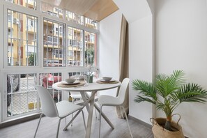 Een eethoek met een ronde tafel, witte stoelen, een potplant en uitzicht op een gebouw door grote ramen.