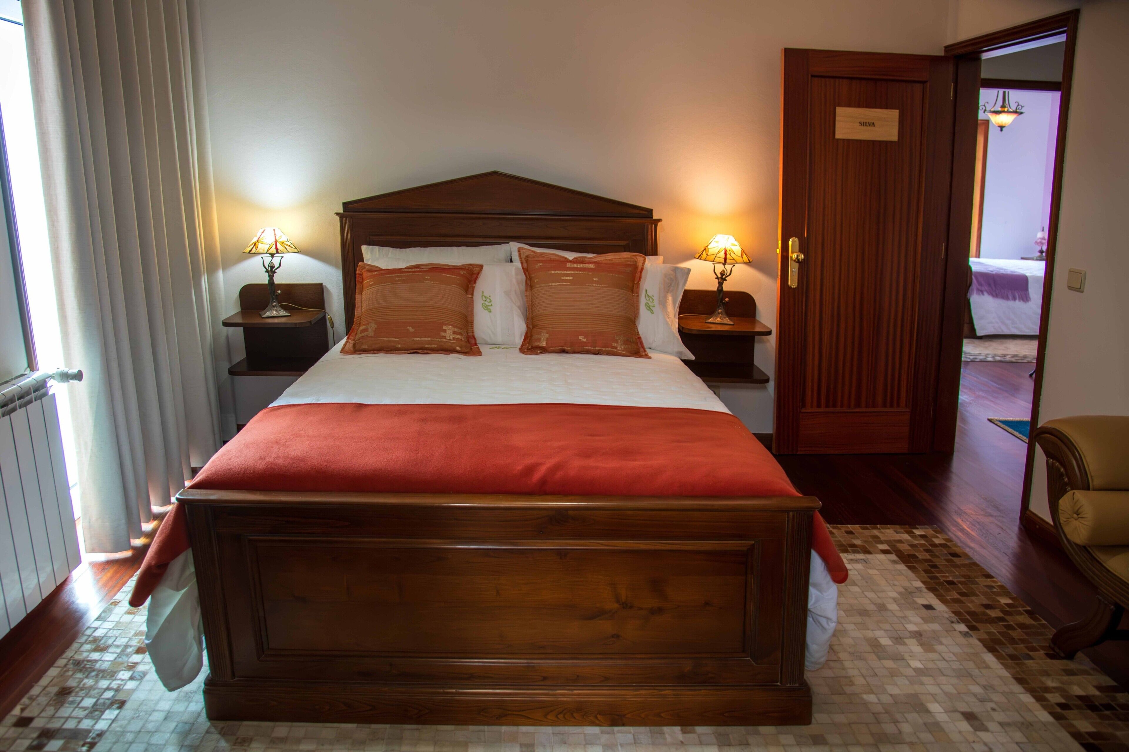 Uma cama grande com cabeceira de madeira, duas mesas de cabeceira com luminárias, uma placa na porta e vista para um corredor.