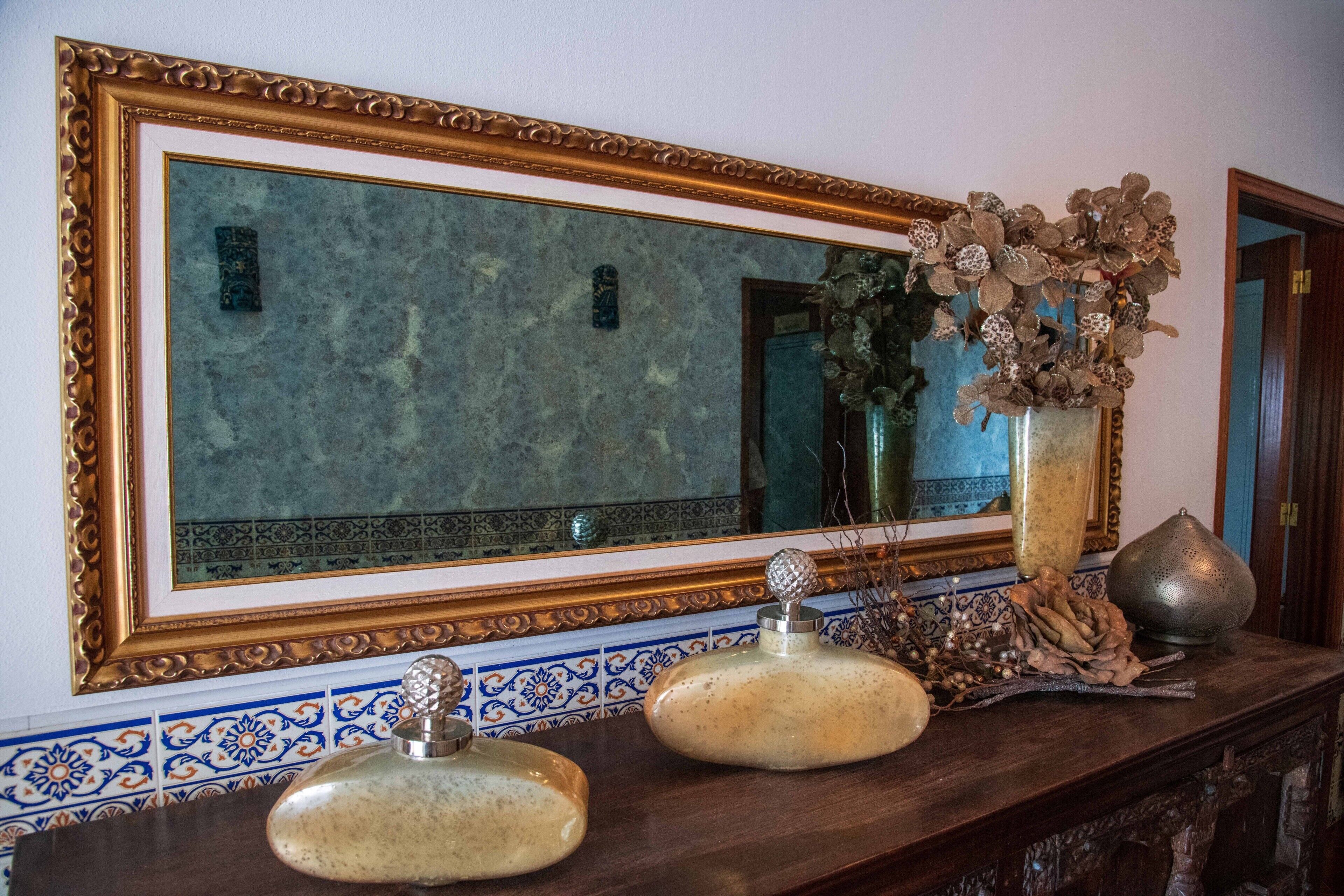 Un espejo con un marco decorativo, un florero con flores secas y dos recipientes de cerámica sobre una superficie de madera.