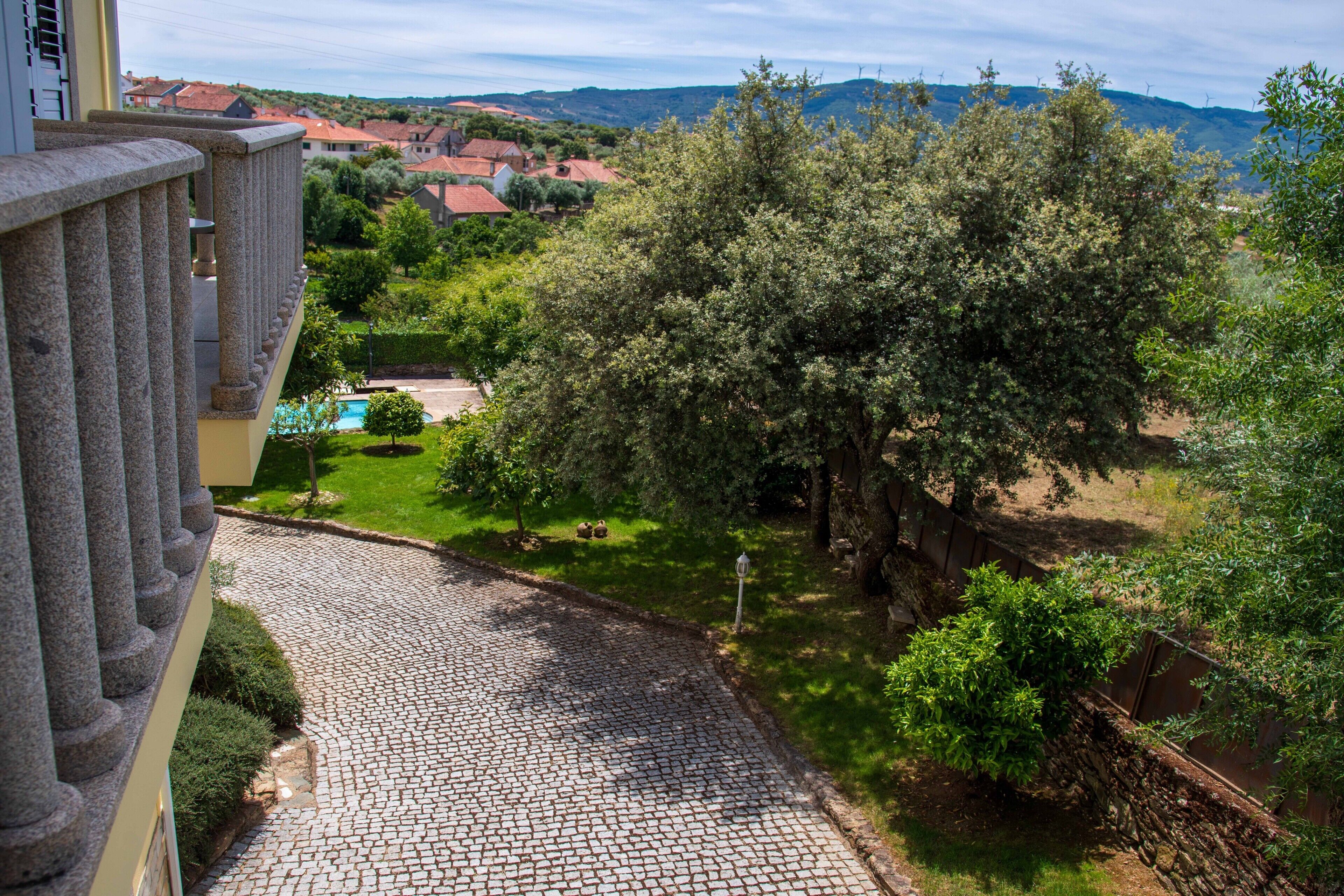 Un sentier pavé de pierres mène à une cour intérieure avec une piscine, entourée d’arbres et offrant une vue sur un village aux maisons aux toits rouges.