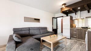 Un salon moderne avec un canapé d’angle gris, une table basse en bois et un coin cuisine doté de meubles sombres et d’un réfrigérateur blanc.