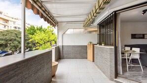 Un balcon avec un sol carrelé, un banc en bois, un pot de fleurs et une vue sur un arbre et un immeuble.