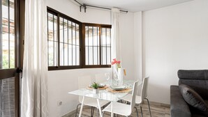 Un coin repas avec une table en verre, des chaises blanches, un vase de fleurs et une vue par une fenêtre ornée de rideaux.