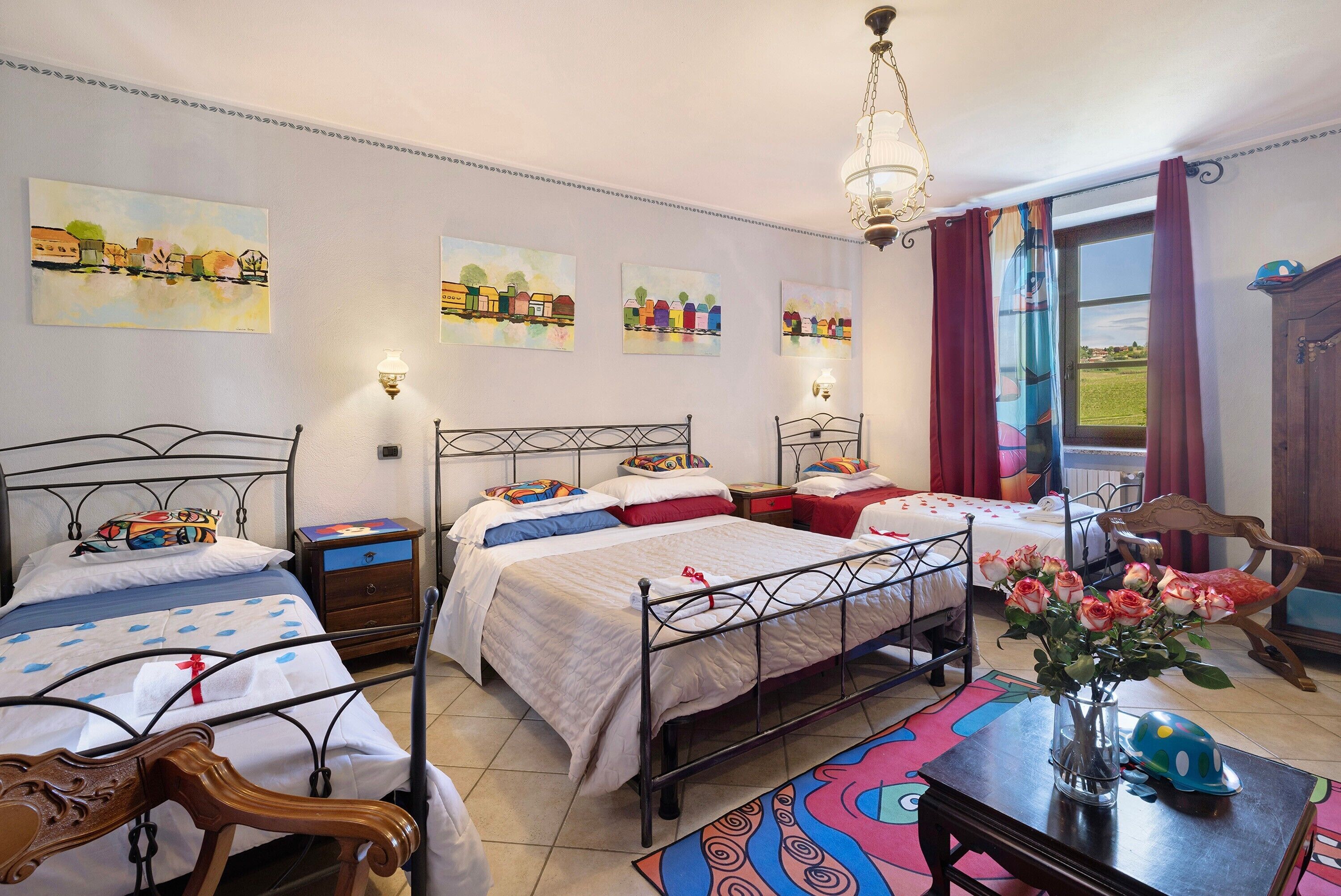 A bedroom with two beds, a wooden chair, a table with a vase of flowers, and a window with curtains.
