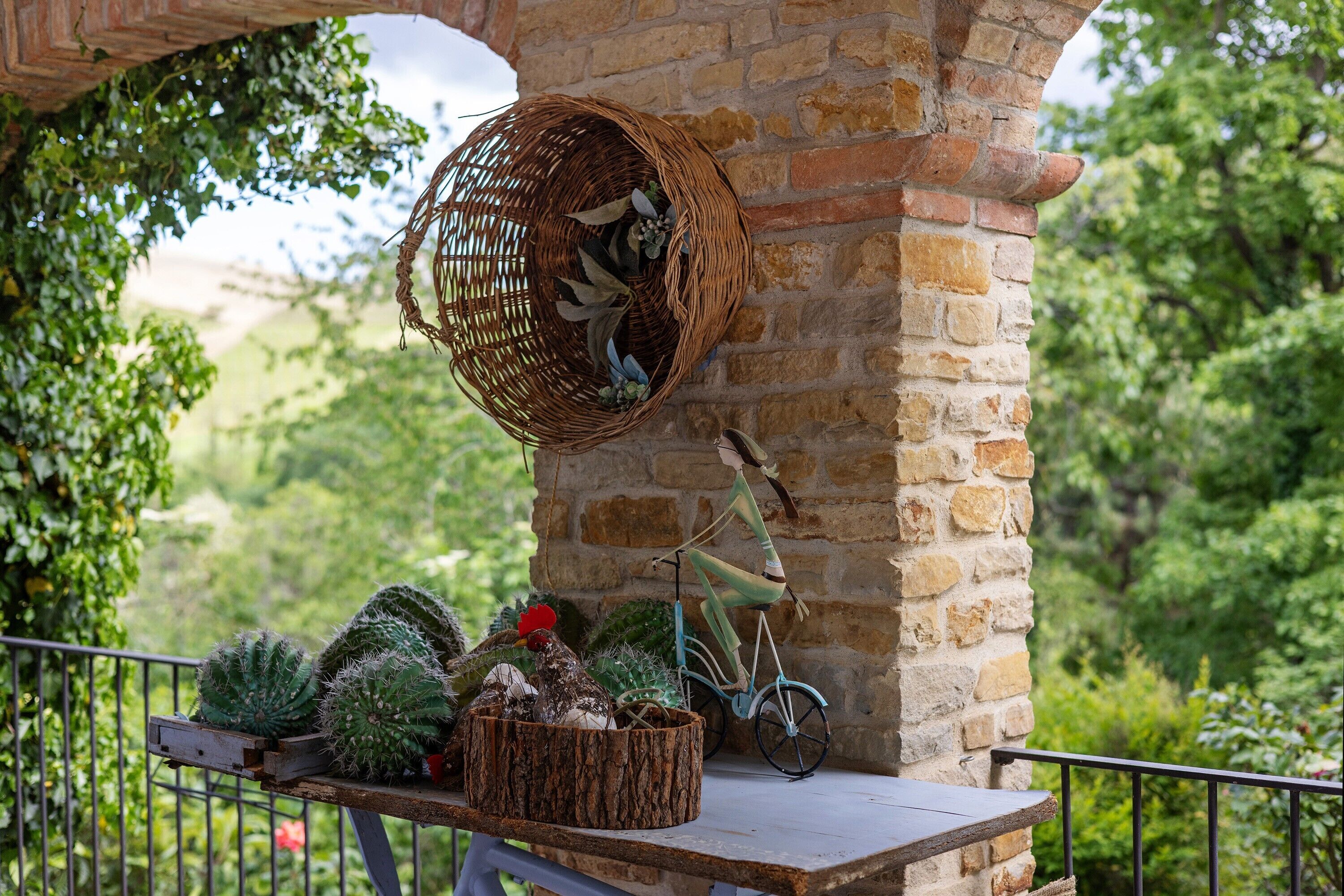 A balcony with a wicker basket, cacti, a bicycle, and a rustic brick wall.