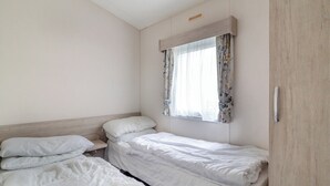 A small, clean room with two single beds, a window with floral curtains, and a wooden wardrobe. - Castaways Oyster 8 Sea View (Pet) (Bacton-on-Sea)