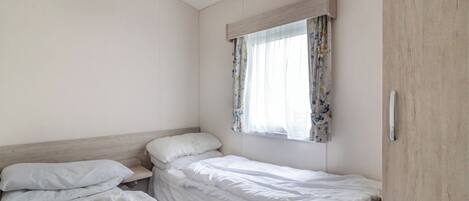 A small, clean room with two single beds, a window with floral curtains, and a wooden wardrobe.