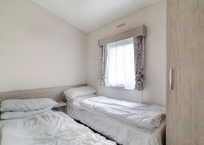 A small, clean room with two single beds, a window with floral curtains, and a wooden wardrobe. - Castaways Oyster 8 Sea View (Pet) (Bacton-on-Sea)