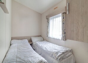 A room with two single beds, a window with floral curtains, a wooden headboard, and a small shelf.