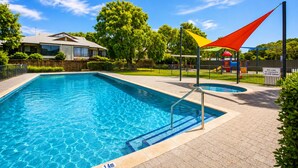 Una piscina con indicador de profundidad de 1,4 metros, rodeada por una zona vallada con un parque infantil y un cartel que indica que no está permitida su uso durante la noche.