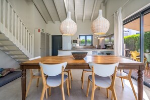 Zona pranzo con tavolo in legno e sedie bianche, cucina sullo sfondo e una scala con ringhiera.