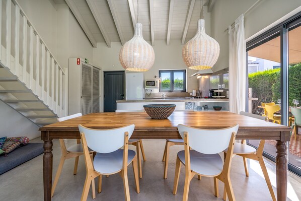 A dining area with a wooden table and white chairs, a kitchen in the background, and a staircase with a railing.