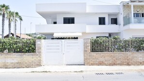 A modern two-story building with a white facade, a gated entrance, and a brick wall with greenery.