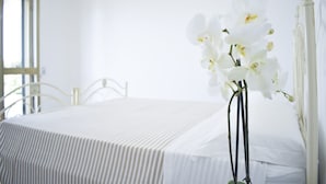 A bed with a white bedspread, a white metal headboard, and a vase with white flowers.