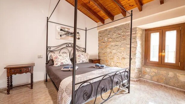 Una cama con dosel sobre un muro de piedra y techo de madera, una mesita de noche pequeña y una ventana con contraventanas de madera.