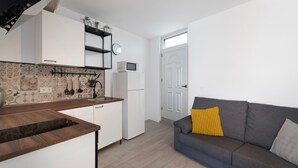 Eine kompakte Küche mit weißen Schränken, einer Mikrowelle und einem Kühlschrank. Ein graues Sofa mit einem gelben Kissen und einer grauen Sitzkissen.