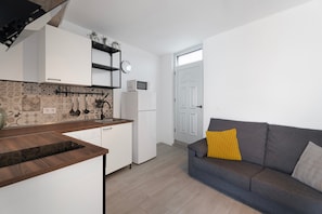 Eine kompakte Küche mit weißen Schränken, einer Mikrowelle und einem Kühlschrank. Ein graues Sofa mit einem gelben Kissen und einer grauen Sitzkissen.
