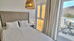 Ein Schlafzimmer mit einem großen Bett, einer Hängeleuchte, einem Nachttisch und Blick ins Freie.