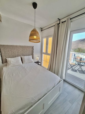 A bedroom with a large bed, a hanging light fixture, a nightstand, and a view of the outdoors. - Apartment 'Apartamento La Pista Sur' with Sea View, Private Terrace and Wi-Fi (San Sebastian de la Gomera)