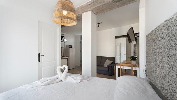 Ein Schlafzimmer mit einem Bett, einer gewobenen Leuchte, einer strukturierten grauen Wand, einem kleinen Tisch mit einer Pflanze und einer Tür zu einer Küchenzeile.