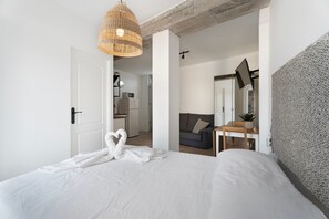Un dormitorio con cama, una lámpara de fibra natural, pared texturizada de color gris, una mesita con planta y un acceso a un kitchenette.
