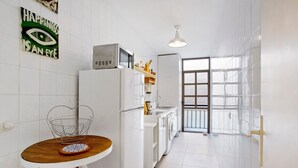 A narrow kitchen with white appliances, a heart-shaped wire basket on a wooden table, and a view through a window to the outside. - Vacation Home 'Casa Brando' with Balcony and Wi-Fi (Corralejo)