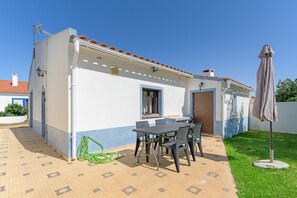 Een huis met een pannendak, een terras met een tafel en stoelen, en een groene tuinslang op de grond.