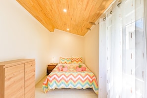 Une chambre avec un plafond en bois, un lit recouvert d’une couverture à motifs zigzag multicolores, une table de chevet avec une lampe et une armoire en bois.