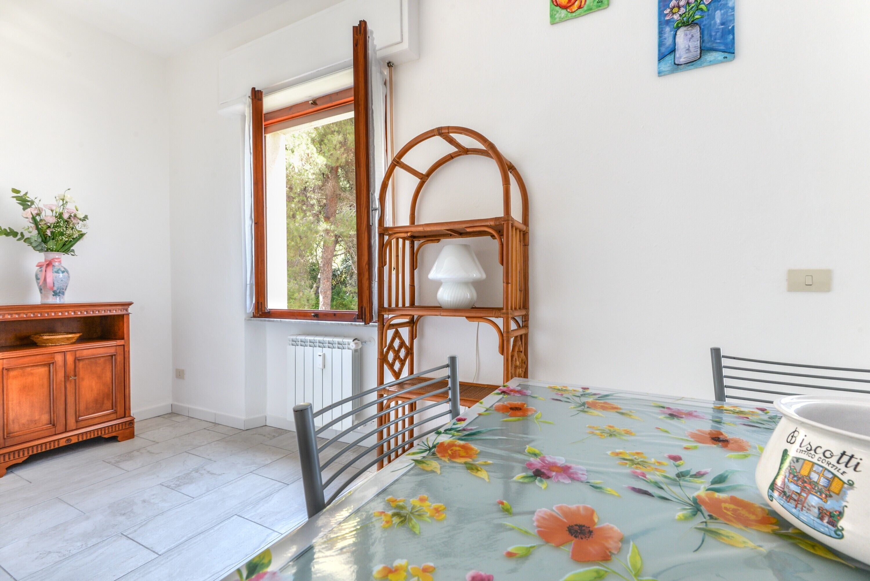 Un coin repas avec une table recouverte d’une nappe à motifs floraux, une chaise en osier, un meuble en bois orné d’un vase de fleurs et une fenêtre donnant sur des arbres.