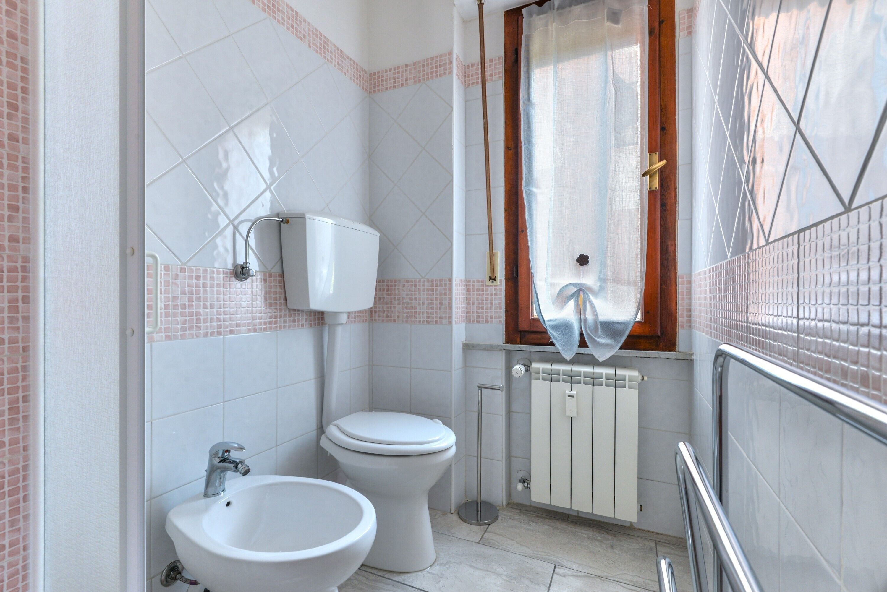 A bathroom with a toilet, bidet, and sink, tiled walls, a radiator, and a window with a curtain.