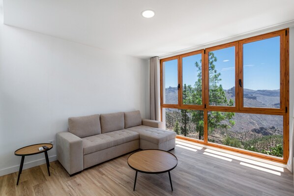 Ein modernes Wohnzimmer mit einem Ecksofa, einem runden Couchtisch und einem großen Fenster mit Blick auf die Berge.