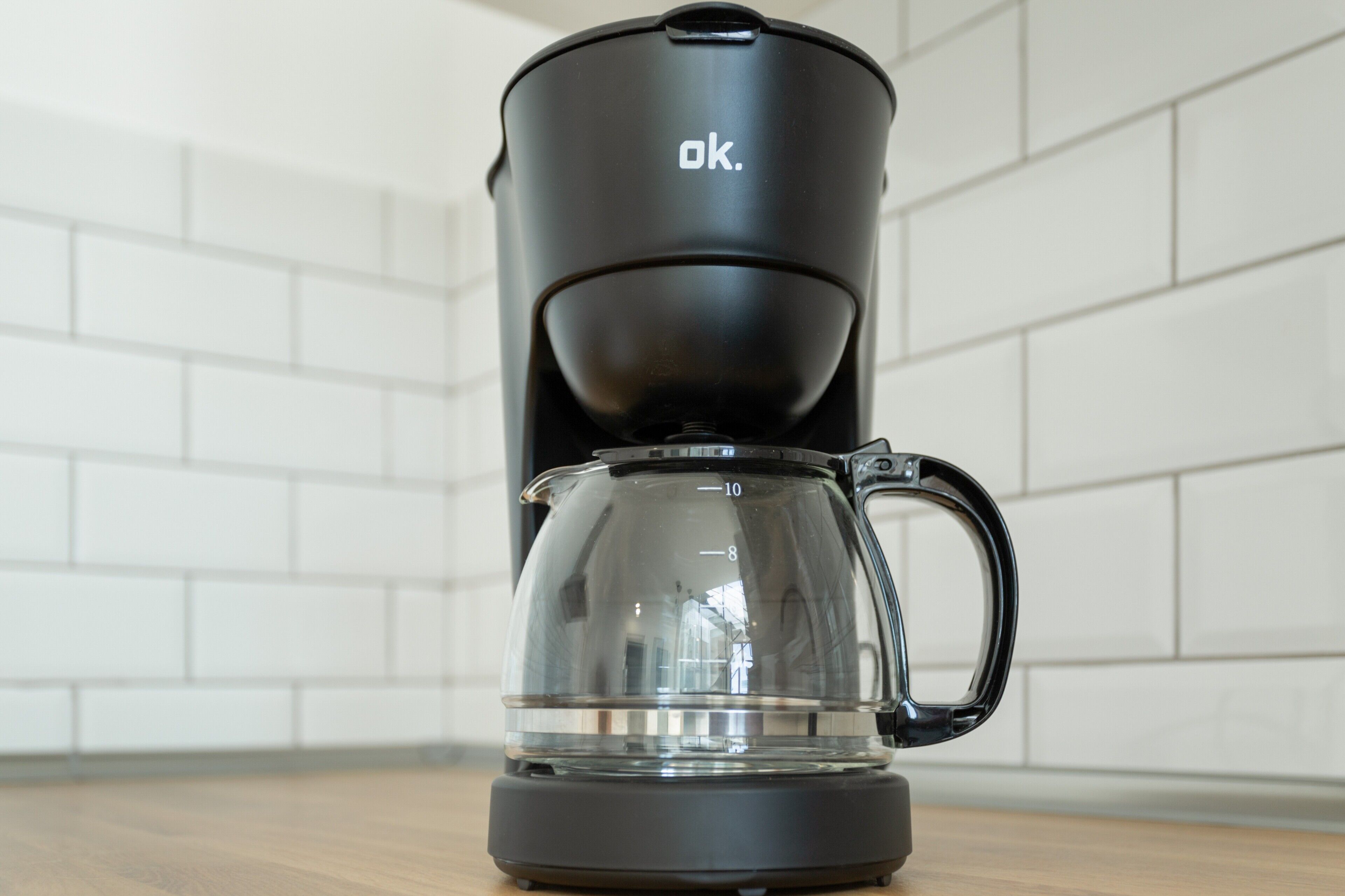 A black coffee maker with a clear glass carafe and a handle, placed on a wooden surface against a white tiled wall.