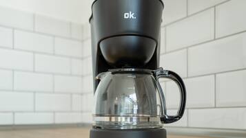 A black coffee maker with a clear glass carafe and a handle, placed on a wooden surface against a white tiled wall.