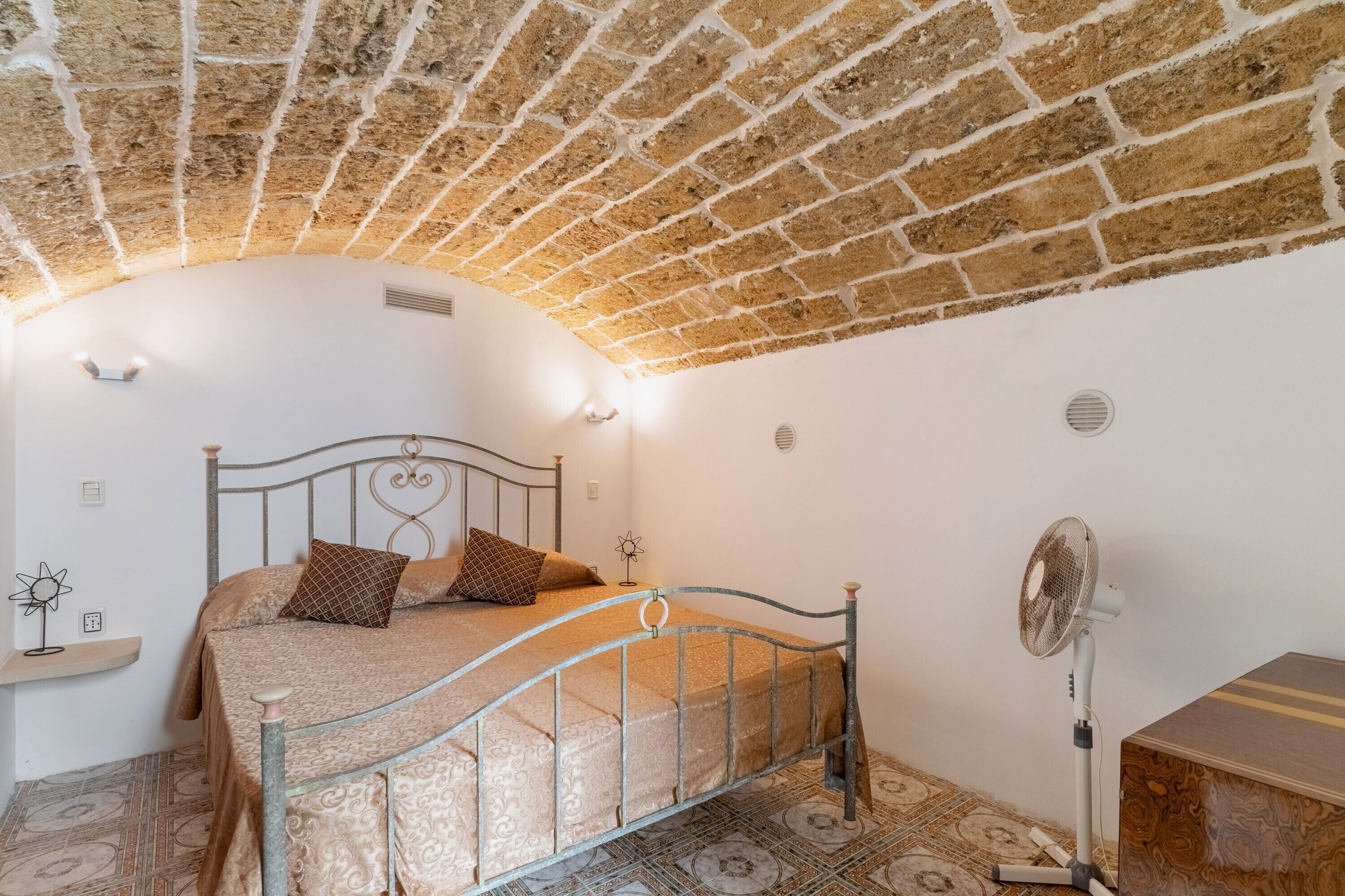 Une chambre avec un plafond en briques, un cadre de lit en métal, une table de chevet, un ventilateur et un revêtement de sol à motifs.