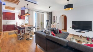 Una sala de estar moderna con un sofá gris, un televisor y una zona de cocina con armarios rojos y una mesa de comedor.