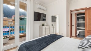 Un dormitorio con cama, televisión, armario y ventana corredera al balcón, con vistas a edificios y montañas.