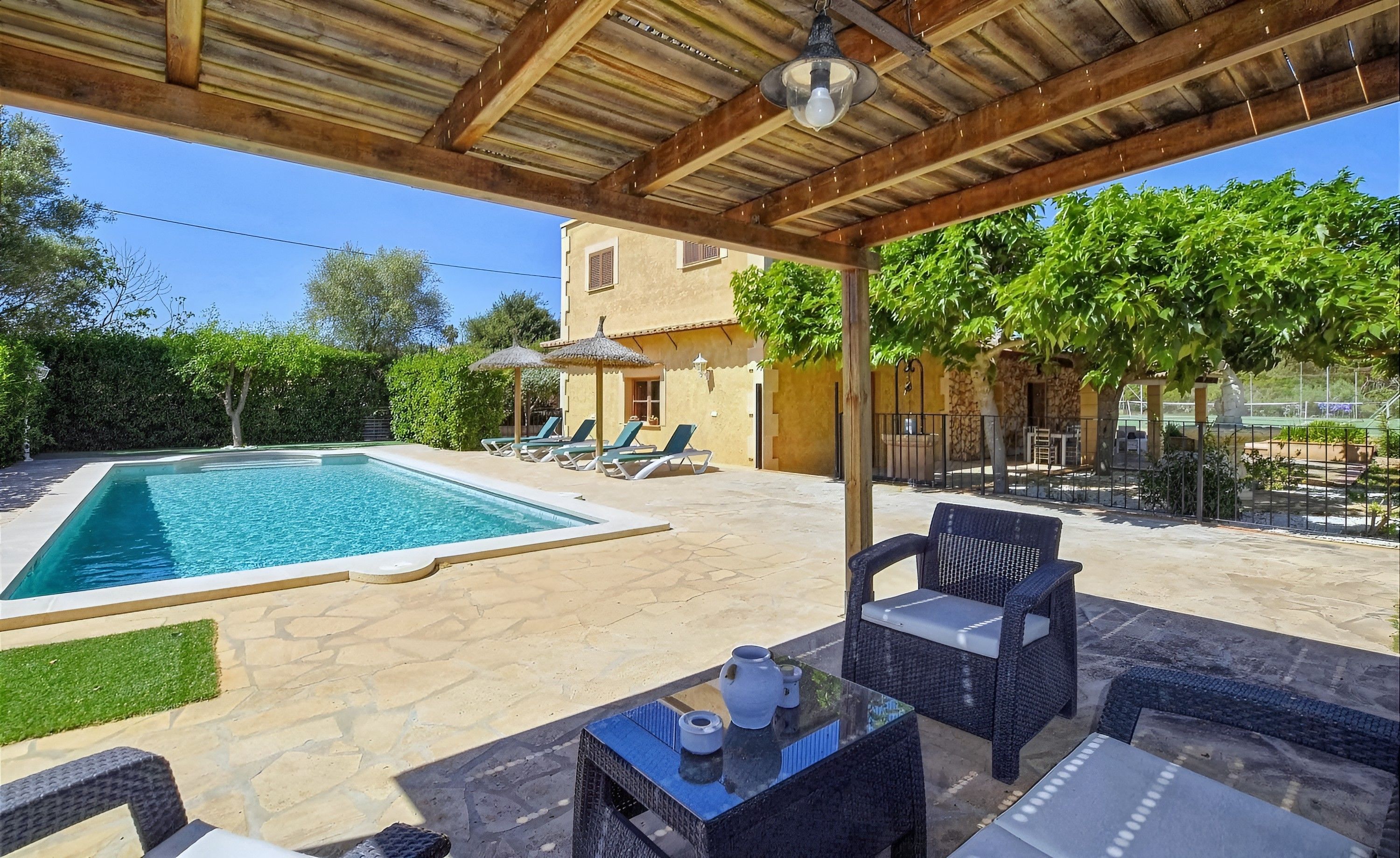 A covered outdoor area with a swimming pool, lounge chairs, and a table with a vase.