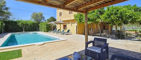 Et overdækket udendørs område med swimmingpool, liggestole og et bord med en vase.