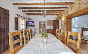 Un espace repas avec une longue table, des chaises en bois, un lustre et un téléviseur fixé au mur.